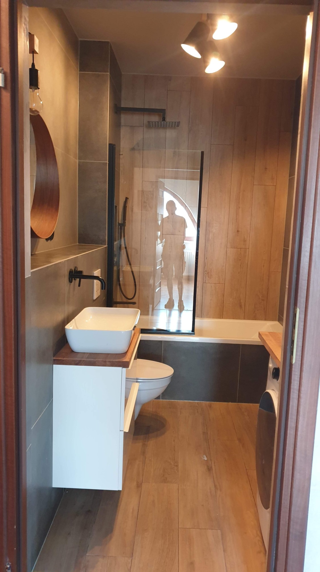 Nowoczesna łazienka z drewnopodobnymi płytkami, kabiną prysznicową typu walk-in z czarnymi profilami, umywalką nablatową na białej szafce i okrągłym lustrem z drewnianą ramą, widoczna sylwetka...