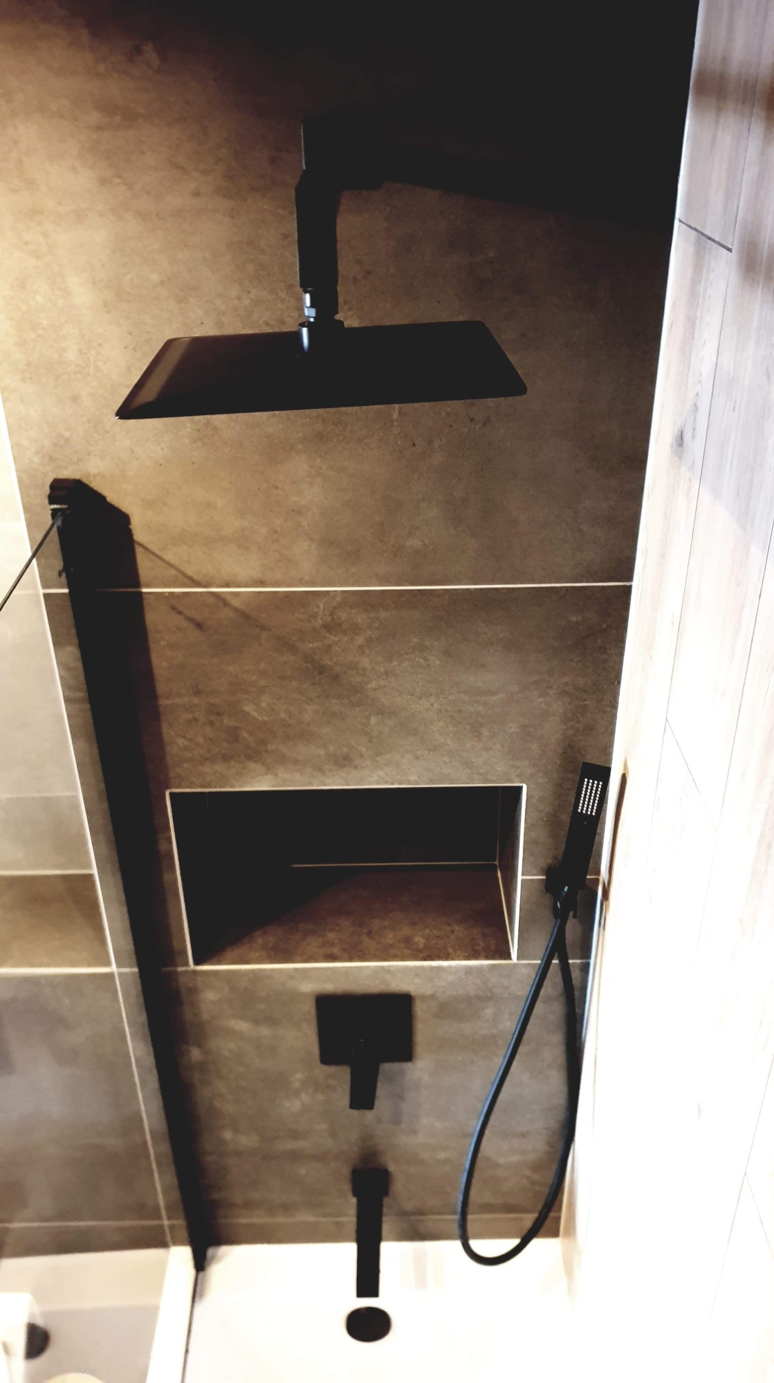 Nowoczesny prysznic z deszczownicą w kolorze czarnym, wbudowana półka w szarej, kamiennej okładzinie, minimalistyczny design.