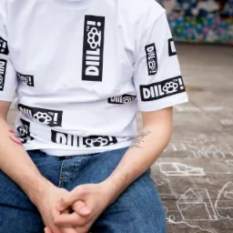 Osoba siedząca na drewnianej powierzchni z rysunkami kredą, ubrana w białą koszulkę z nadrukiem 'DILL!' i symbolem kastetu, widoczny tatuaż na przedramieniu.