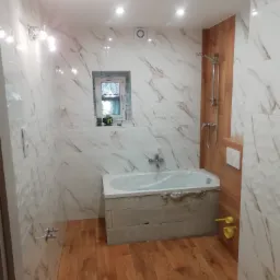 Wnętrze łazienki w trakcie prac wykończeniowych, widoczne płytki imitujące marmur na ścianach, drewnopodobna podłoga, wanna obudowana bloczkami, okno z widokiem na zewnątrz, oraz elementy...