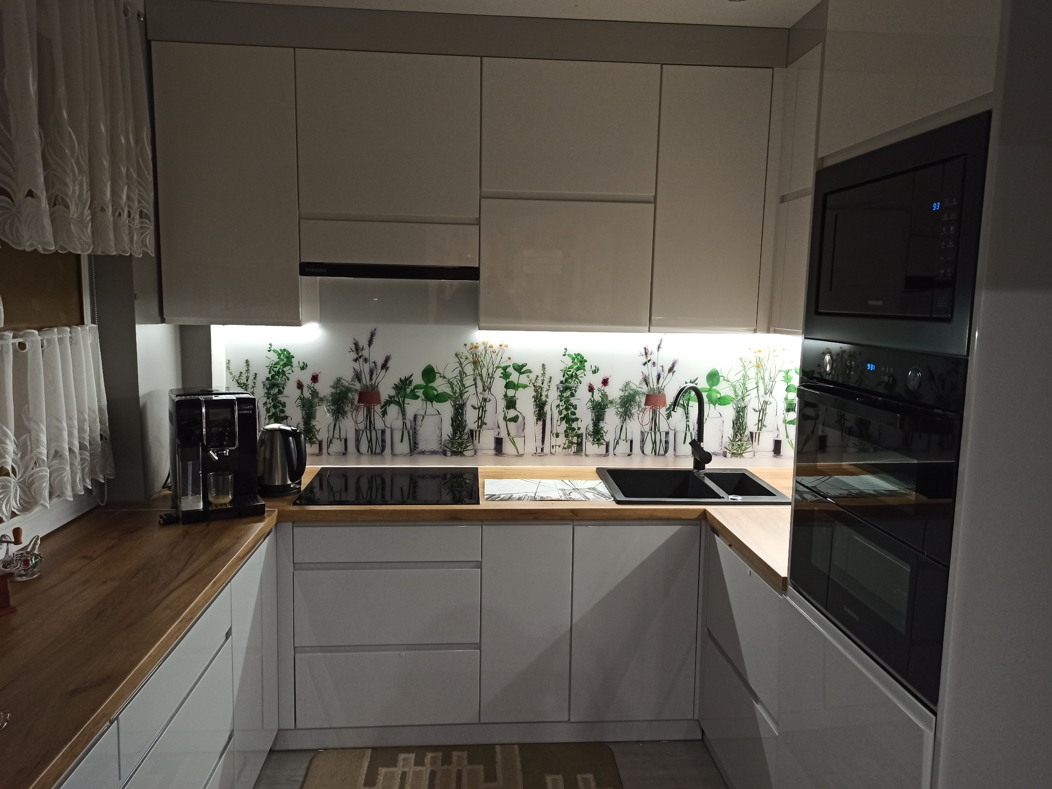 Nowoczesna kuchnia z białymi, lakierowanymi frontami, drewnianym blatem i panelem szklanym z motywem ziół i roślin w szklanych naczyniach, zabudowany piekarnik z mikrofalówką oraz ekspres do kawy.