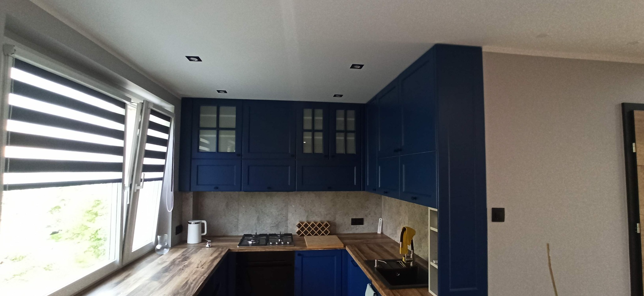 Niebieska zabudowa kuchenna z drewnianym blatem, widok na okno z roletami dzień-noc, płyta gazowa i czarny zlew.