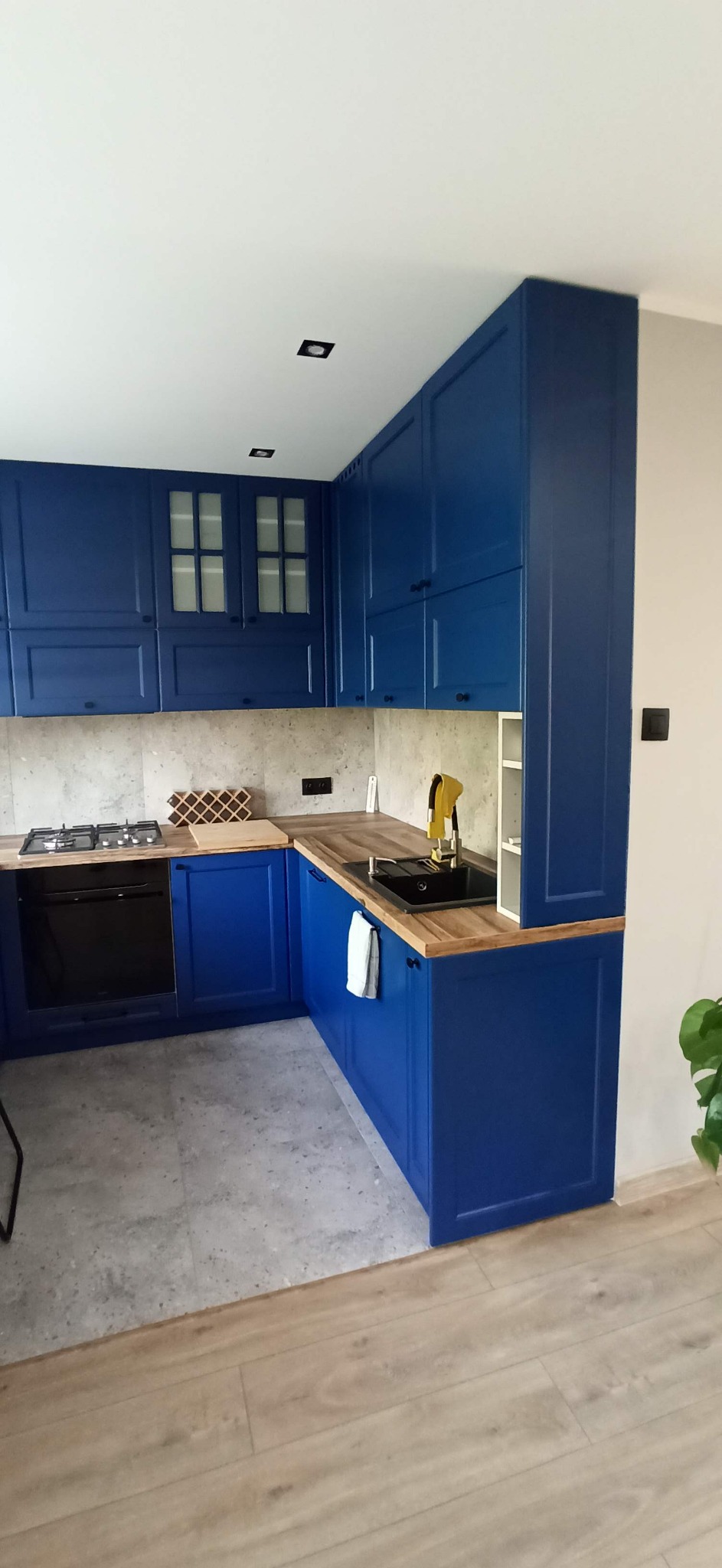 Narożna zabudowa kuchenna w odcieniach granatu z drewnianym blatem, czarnym zlewem i płytą grzewczą, uzupełniona o szklane witryny w górnych szafkach.