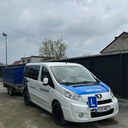 Firma Hanex Plus Elżbieta Kukla - Pojazd szkoły jazdy OSK Hanex z przyczepą, oznaczony niebieskim pasem i literą 'L', zaparkowany na szutrowej nawierzchni przed czarnym płotem.