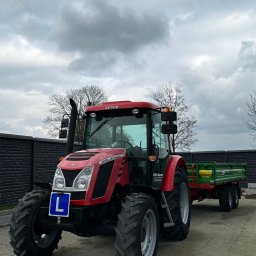 Firma Hanex Plus Elżbieta Kukla - Czerwony ciągnik Zetor z przyczepą i tablicą 'L' na tle pochmurnego nieba i czarnego ogrodzenia.