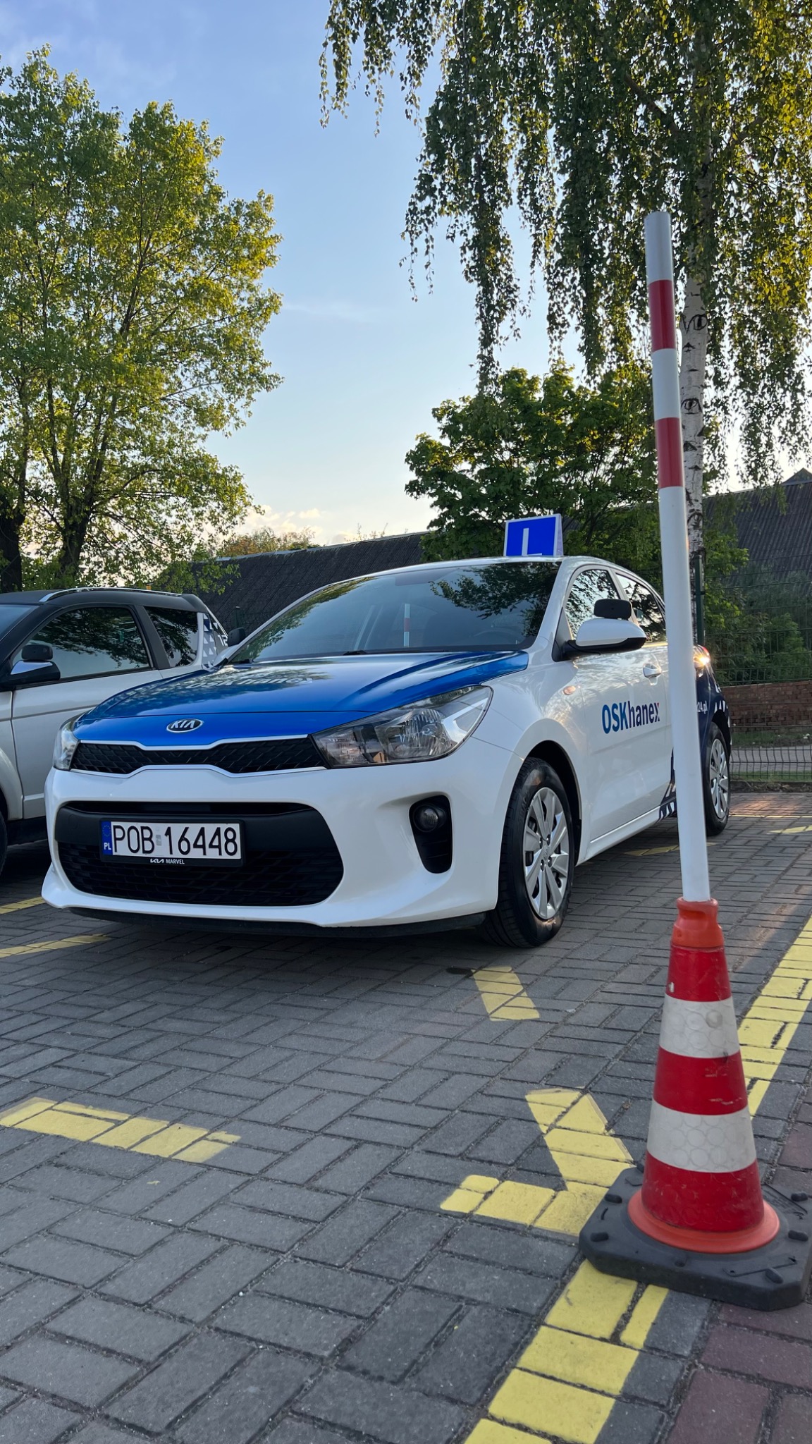 Biały samochód z niebieskim pasem na masce i literą 'L' na dachu, zaparkowany na placu manewrowym z namalowanymi żółtymi liniami, obok czerwono-białego pachołka, w tle drzewa.