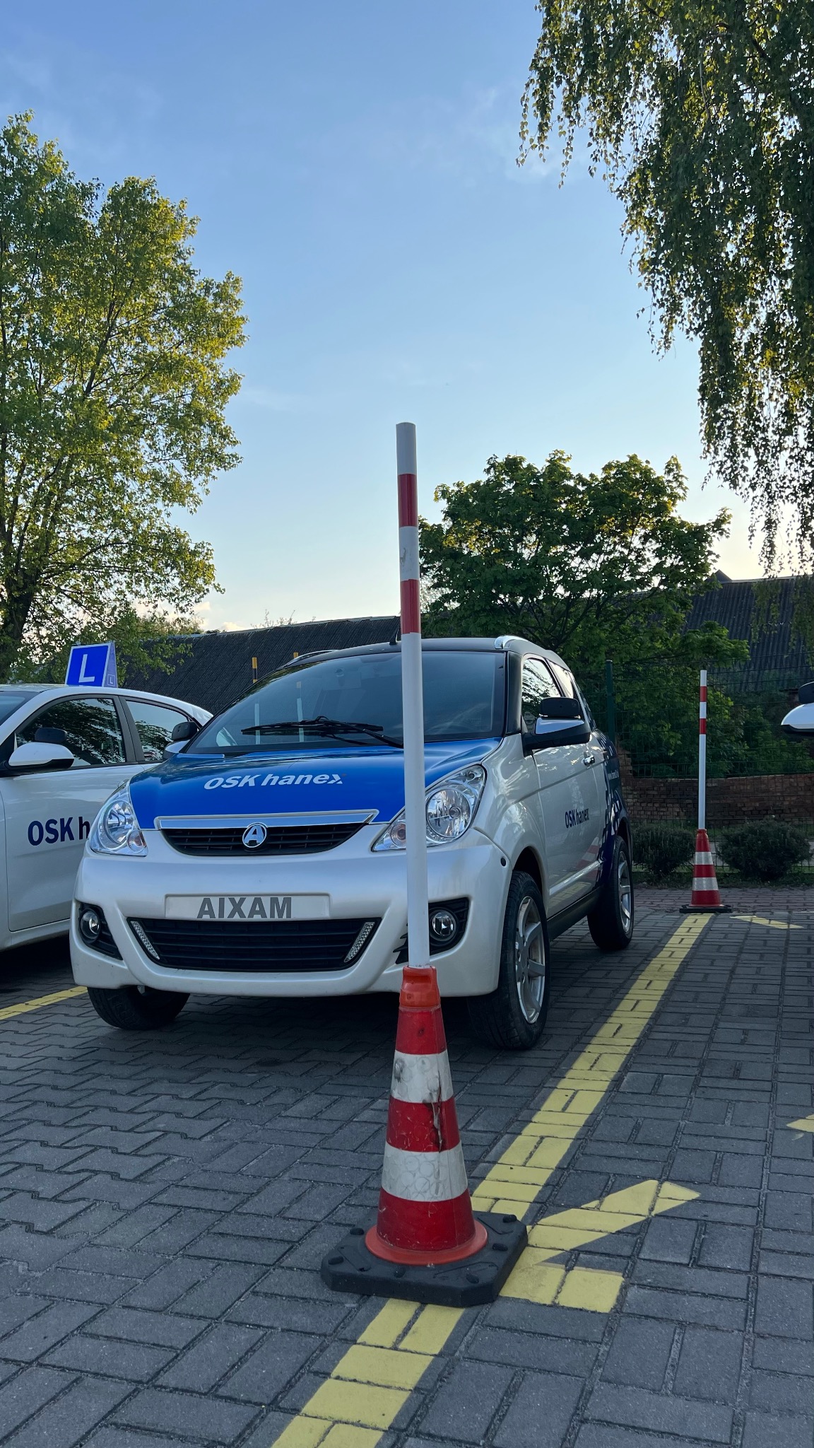 Biały samochód marki Aixam z niebieskim pasem i napisem OSK Hanex, zaparkowany na placu manewrowym z pachołkami i wymalowanymi liniami, w tle zieleń drzew.
