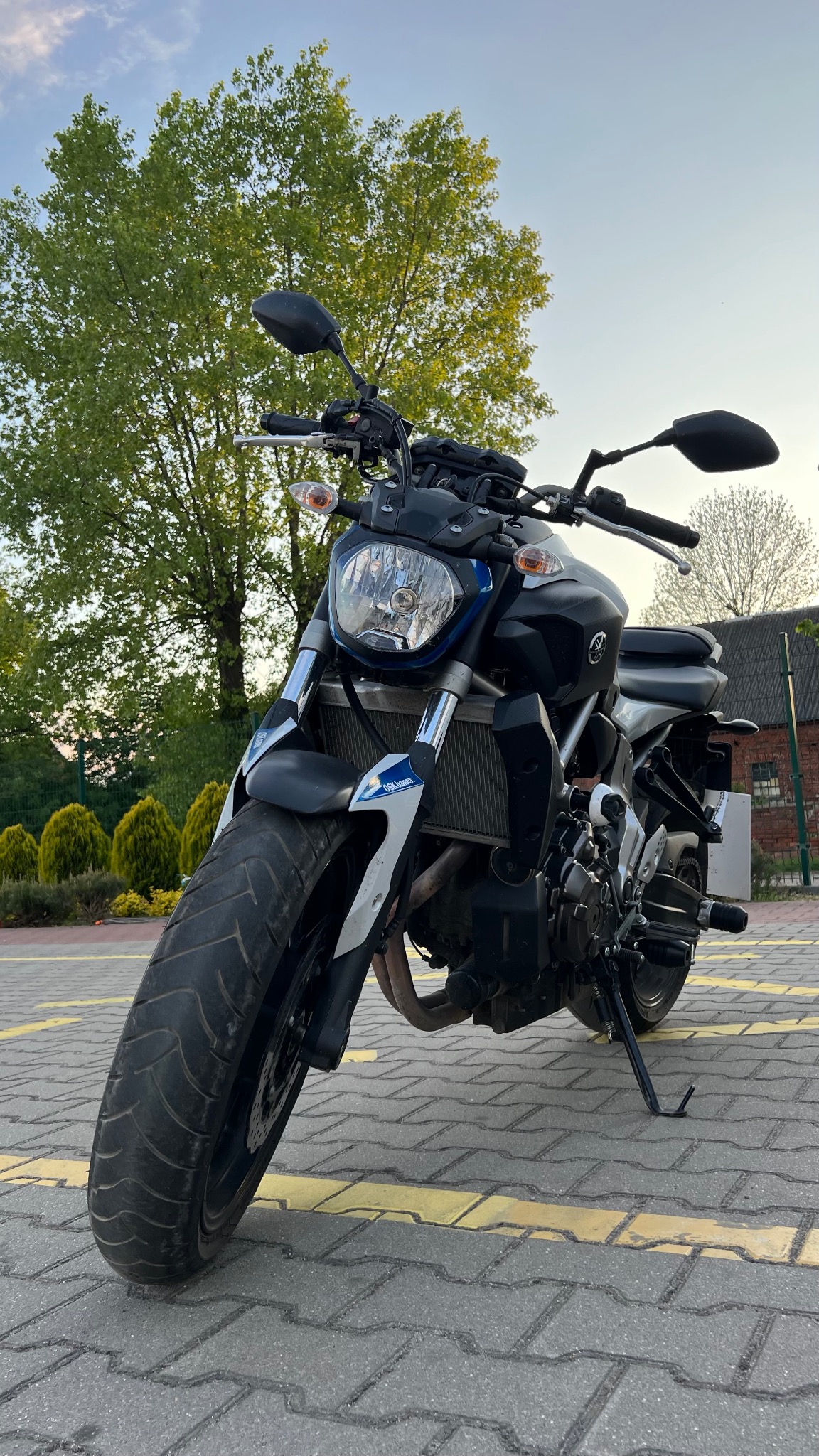 Czarny motocykl Yamaha MT-07 zaparkowany na kostce brukowej z żółtymi liniami, widok z przodu, w tle drzewa i budynek.