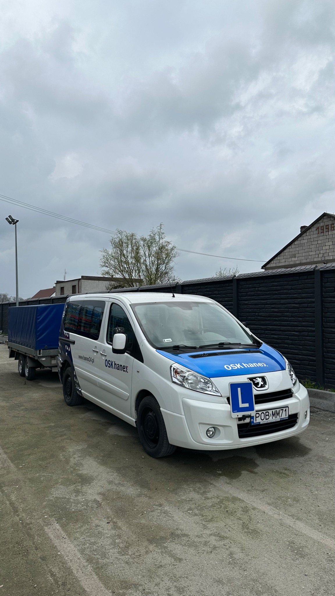 Pojazd szkoły jazdy OSK Hanex z przyczepą, oznaczony niebieskim pasem i literą 'L', zaparkowany na szutrowej nawierzchni przed czarnym płotem.