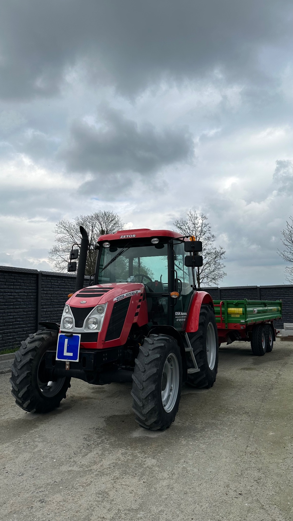 Czerwony ciągnik Zetor z przyczepą i tablicą 'L' na tle pochmurnego nieba i czarnego ogrodzenia.