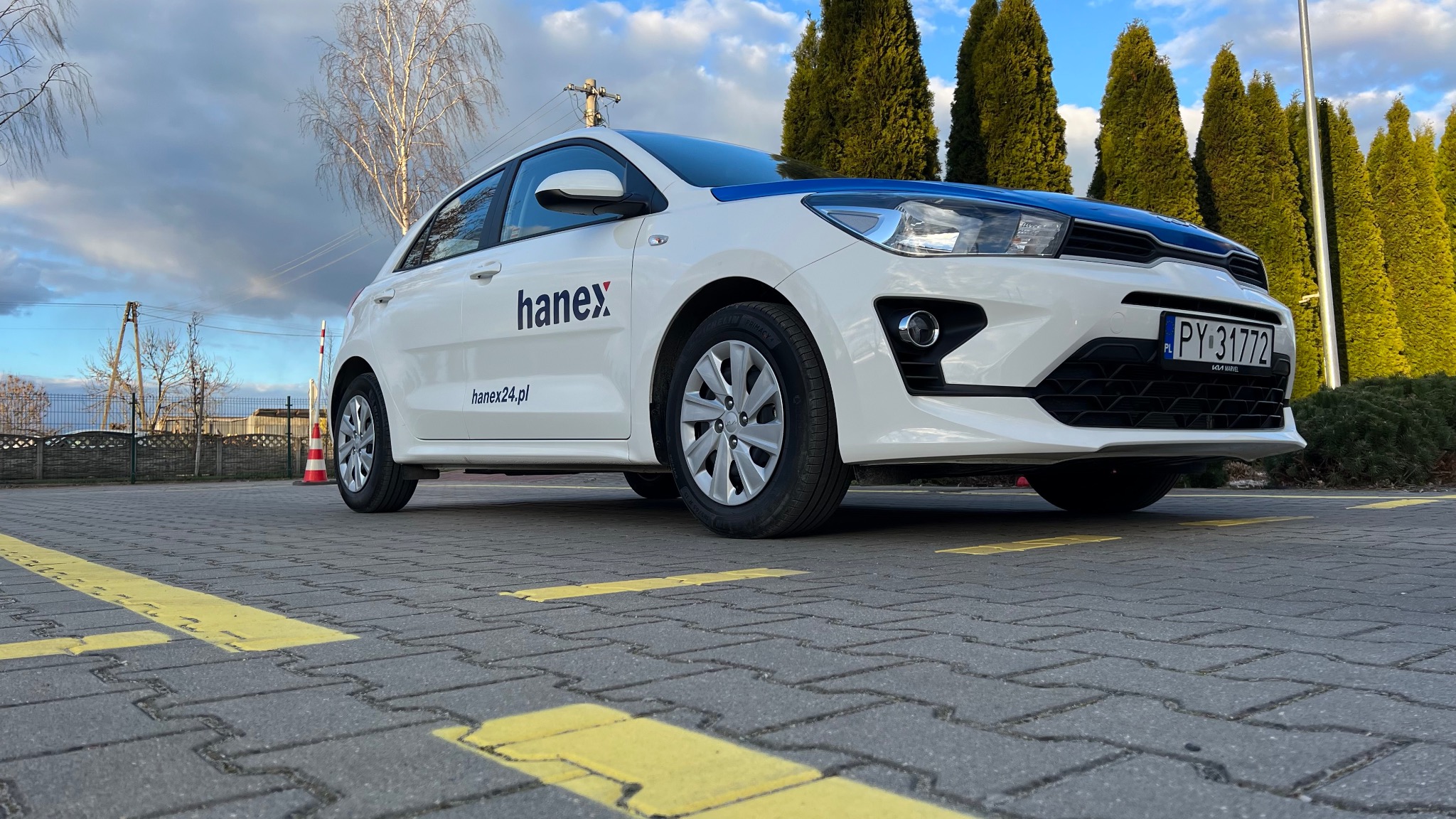 Biały samochód z niebieskim pasem i logo 'hanex' na parkingu z żółtymi liniami, tłem są zielone drzewa i błękitne niebo.