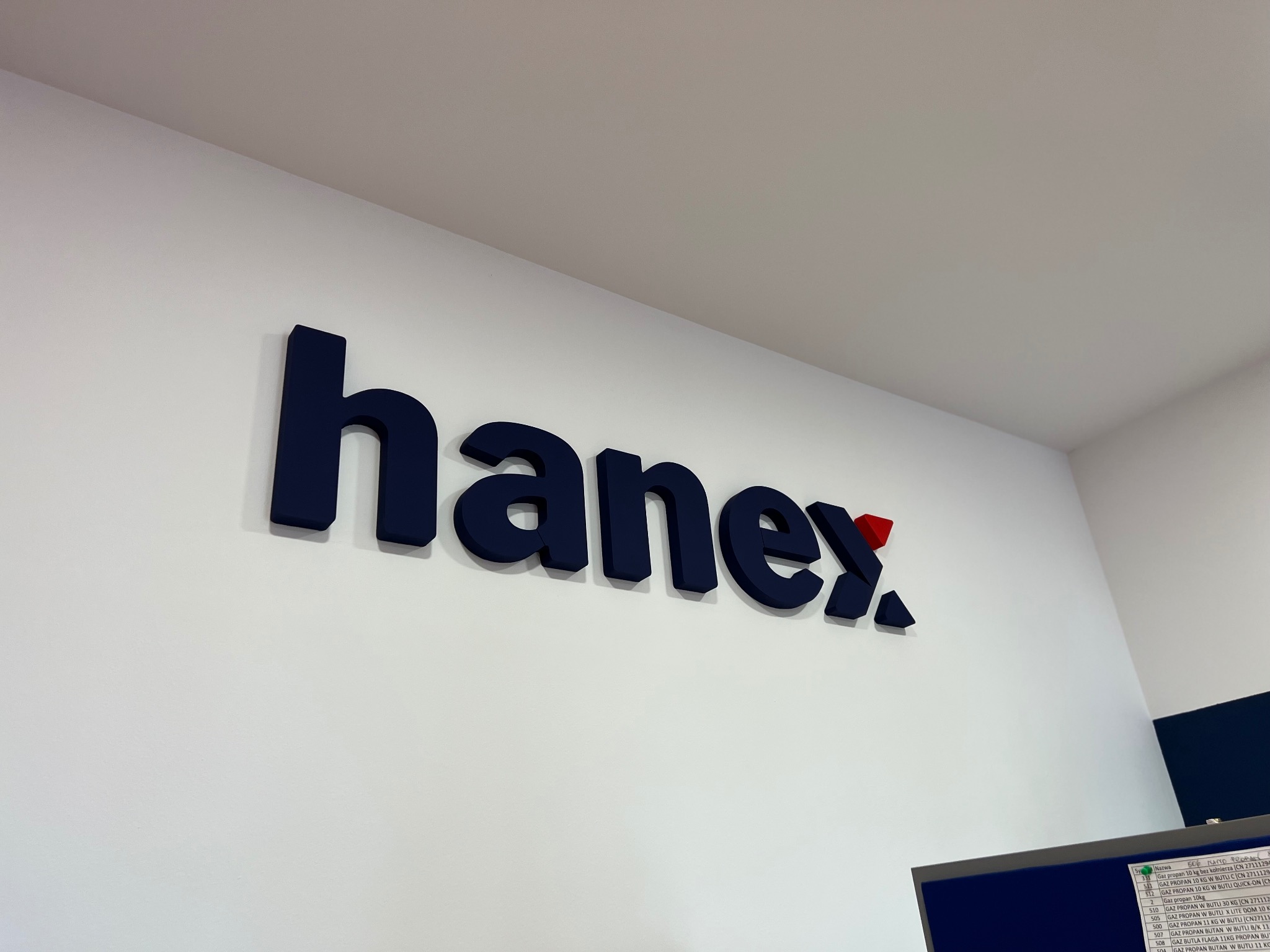 Logo 'hanex.' w granatowym kolorze z czerwonym akcentem na białej ścianie, widoczny fragment monitora z listą pozycji.