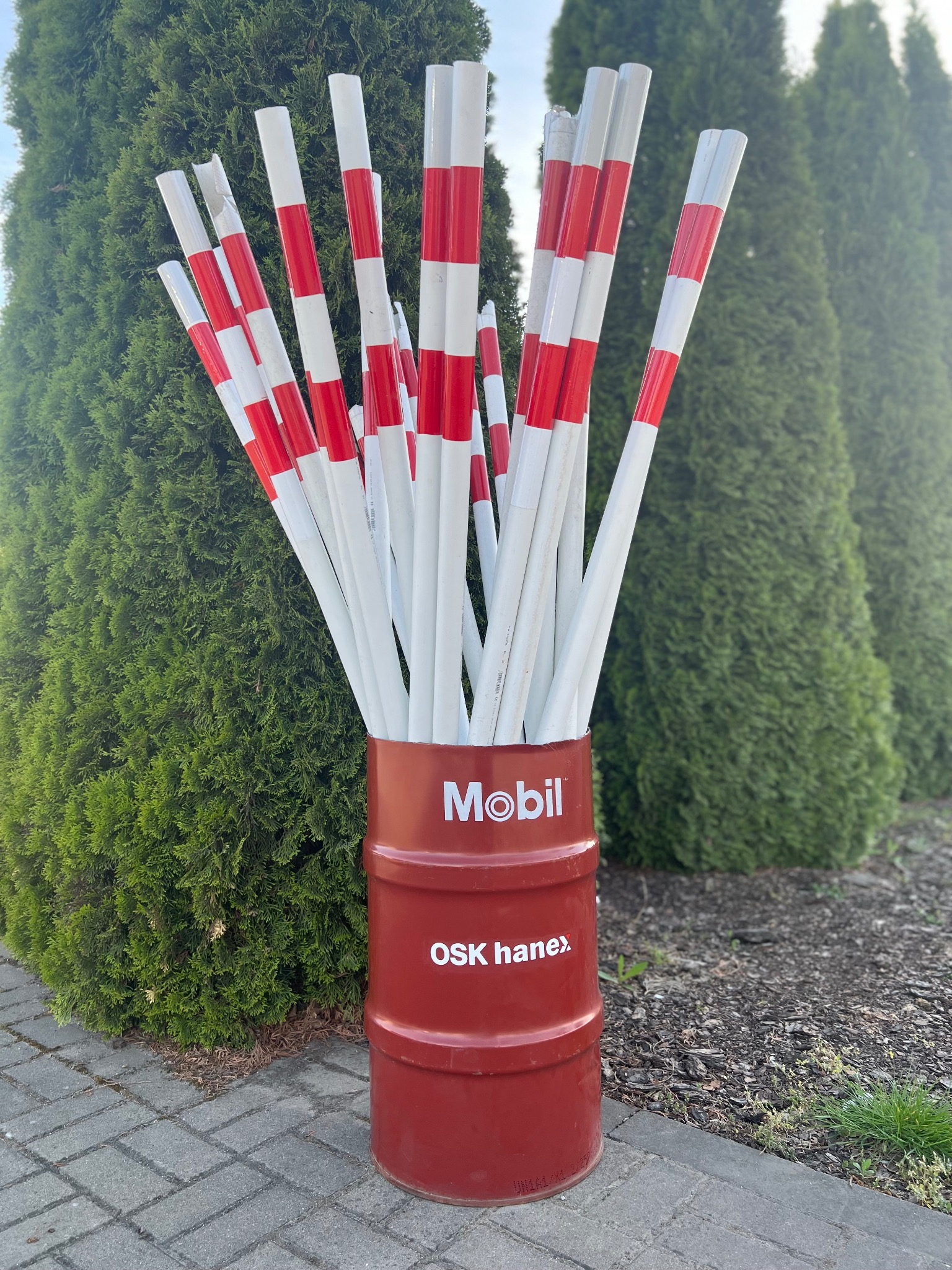 Słupki do nauki jazdy z czerwono-białymi pasami, umieszczone w czerwonej beczce z logo Mobil i nazwą OSK Hanex, na tle zielonych krzewów.