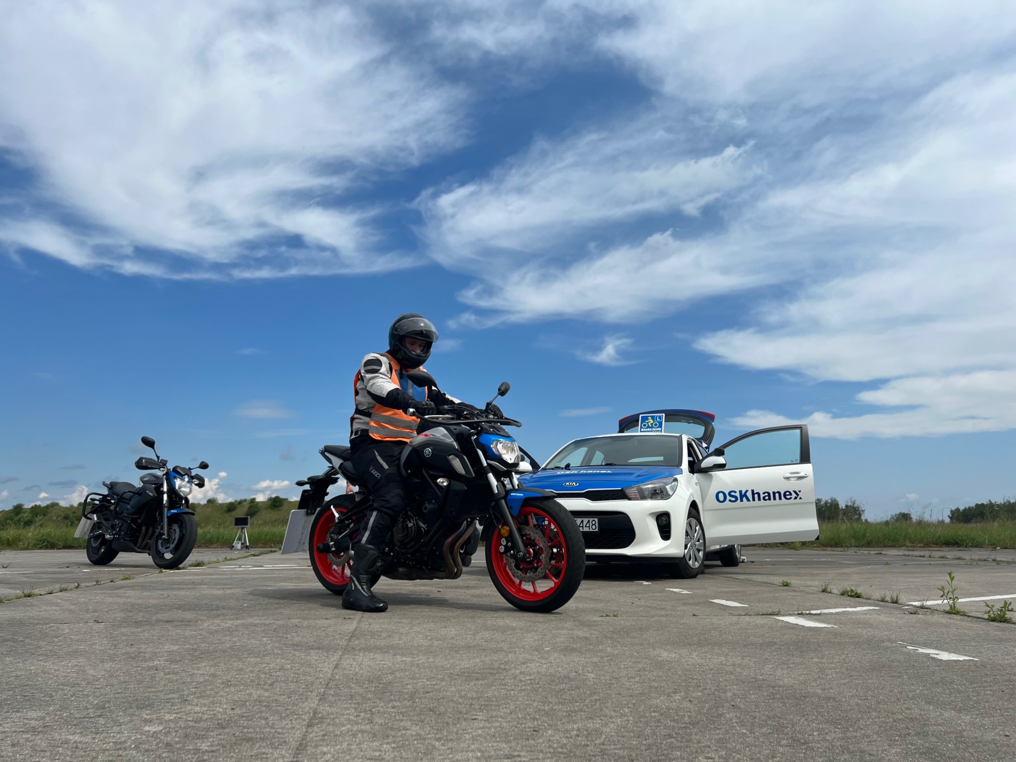 Instruktor szkoły jazdy na motocyklu Yamaha MT-07 z czerwonymi felgami, obok biały samochód Kia Rio z logo OSK Hanex i niebieskim pasem na dachu, w tle drugi motocykl i błękitne niebo z chmurami.