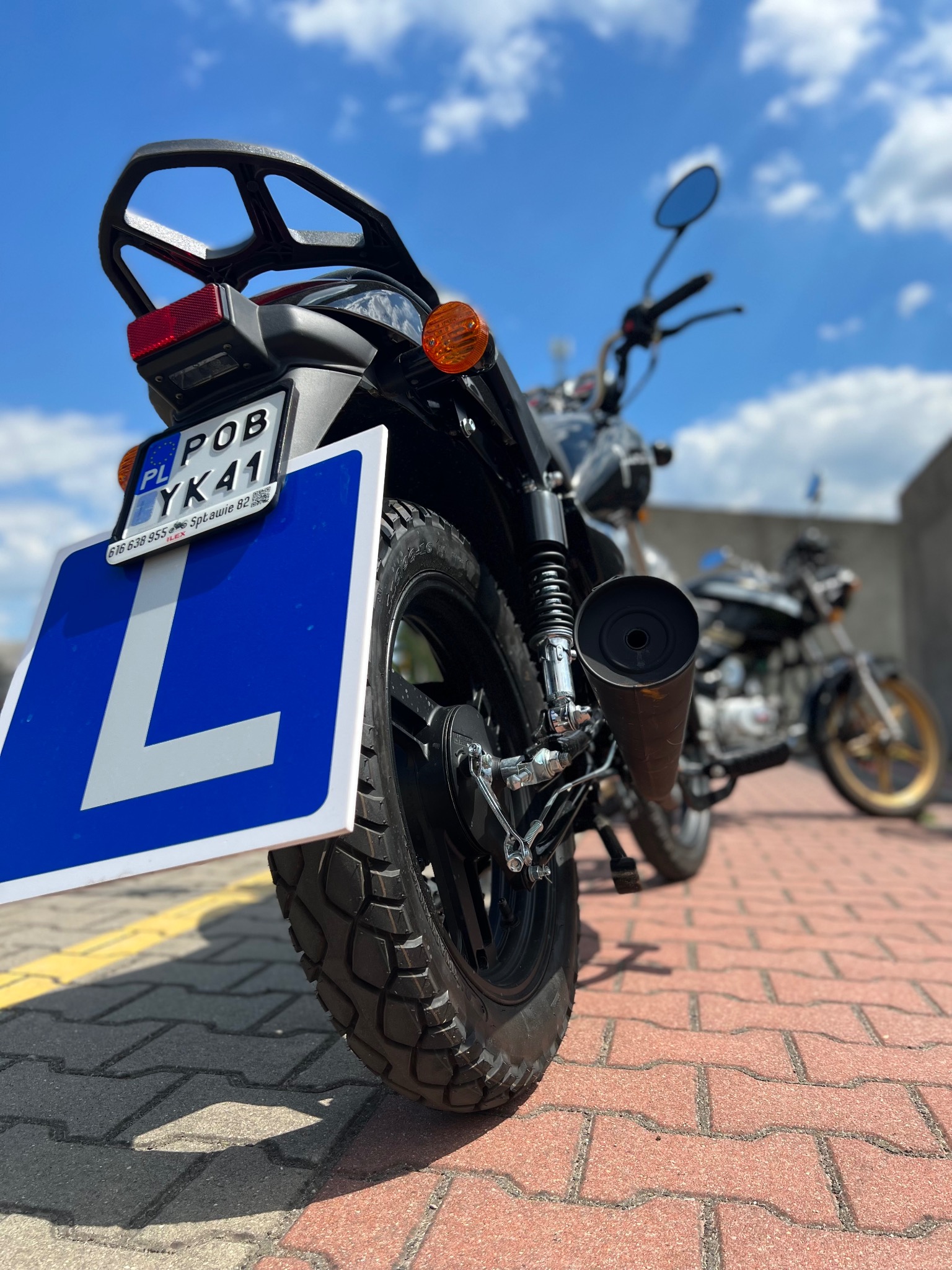 Motocykl z niebieską tablicą z literą L, widok z tyłu, na tle kostki brukowej i drugiego motocykla w oddali.