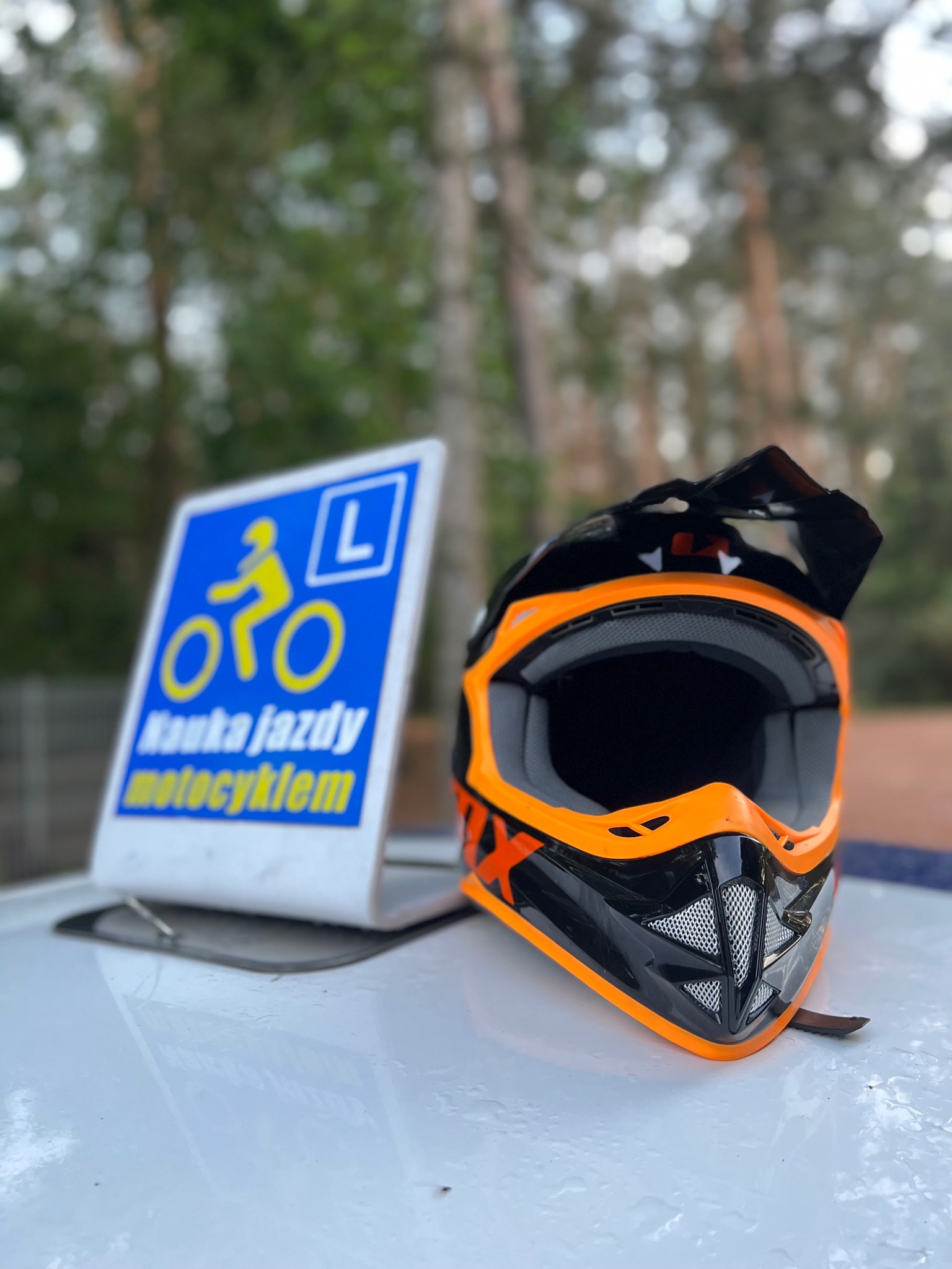 Czarno-pomarańczowy kask motocyklowy obok znaku z symbolem motocyklisty i napisem 'Nauka jazdy motocyklem', w tle las.