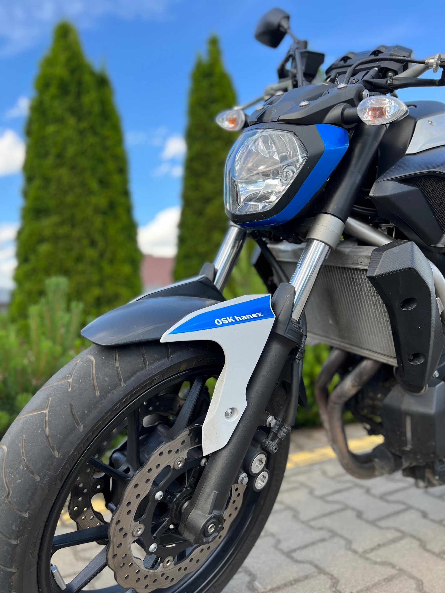 Motocykl z widoczną przednią lampą, oponą i napisem 'OSK Hanex' na błotniku, na tle tuj i błękitnego nieba.