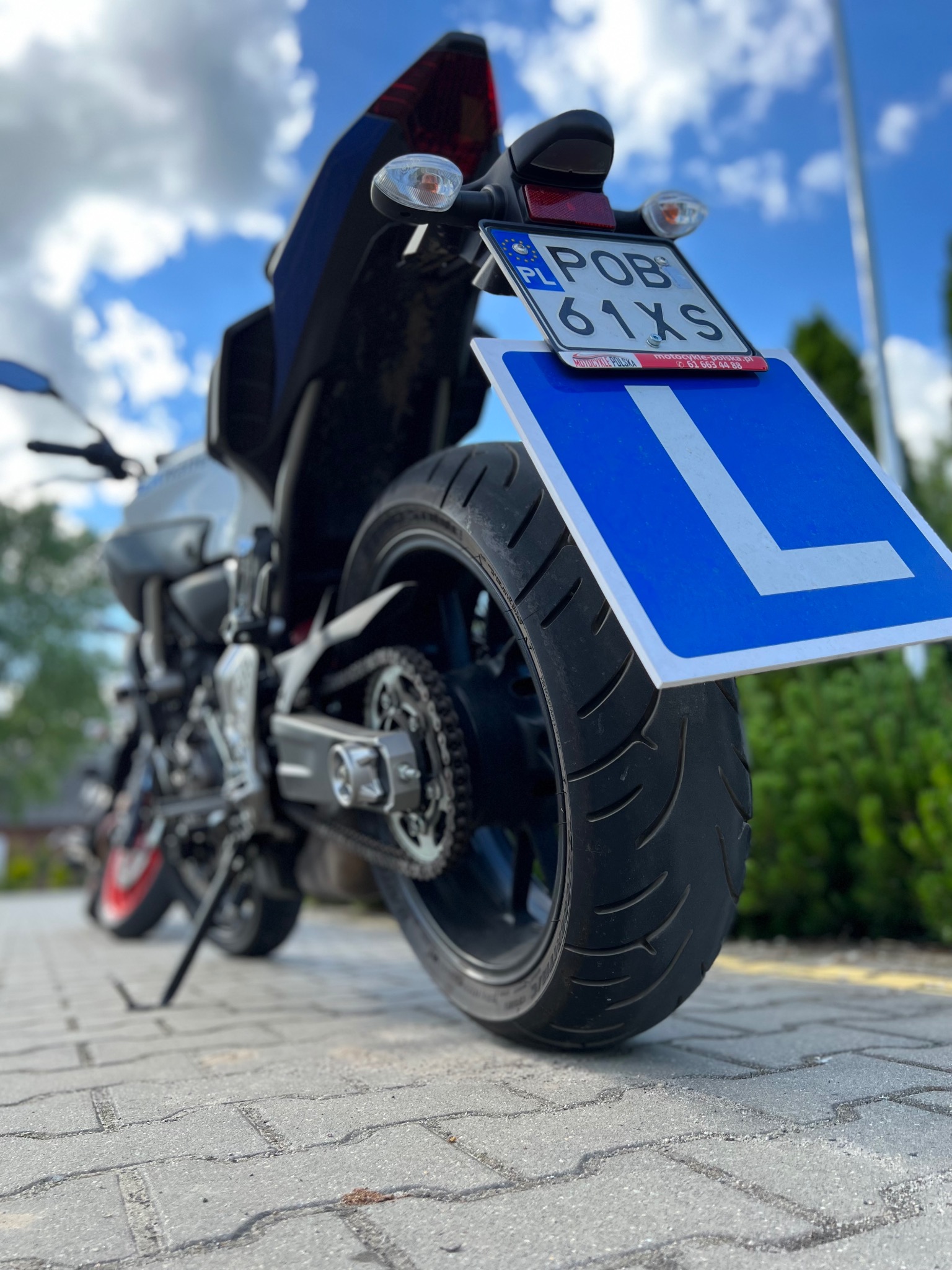 Motocykl z widoczną tablicą rejestracyjną i niebieską literą L, stojący na kostce brukowej, z zielenią i błękitnym niebem w tle.