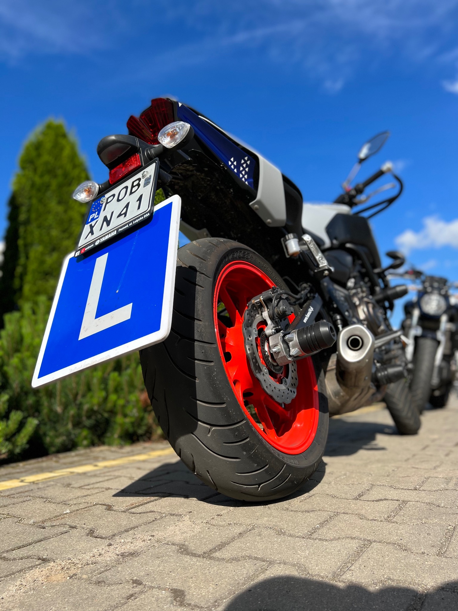 Motocykl z czerwoną felgą i niebieską tablicą z literą L, stojący na brukowanym placu, w tle zieleń i błękitne niebo.