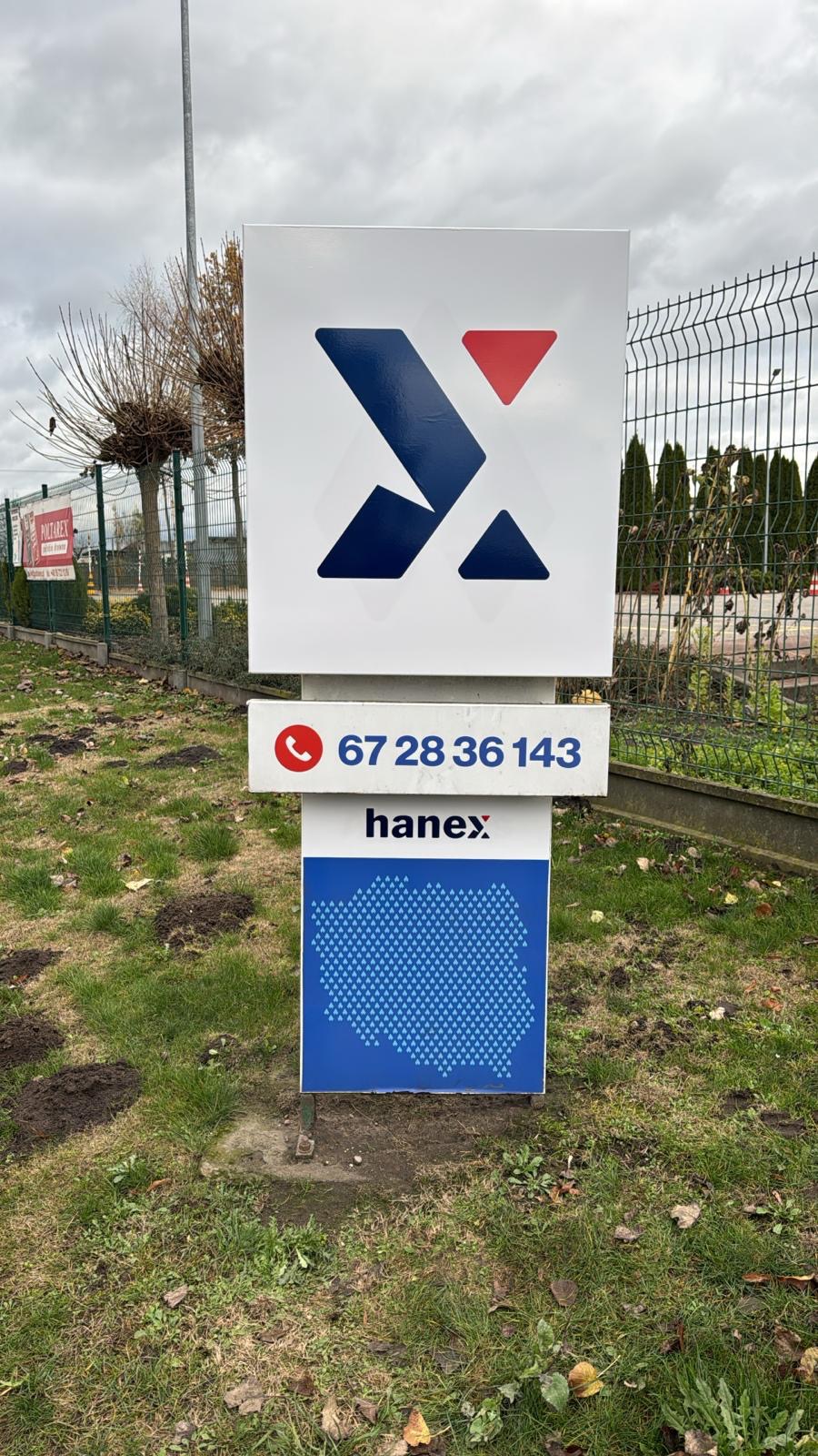 Słup reklamowy z logo firmy Hanex, numerem telefonu i mapą Polski w kropki, na tle trawnika i drzew bez liści.