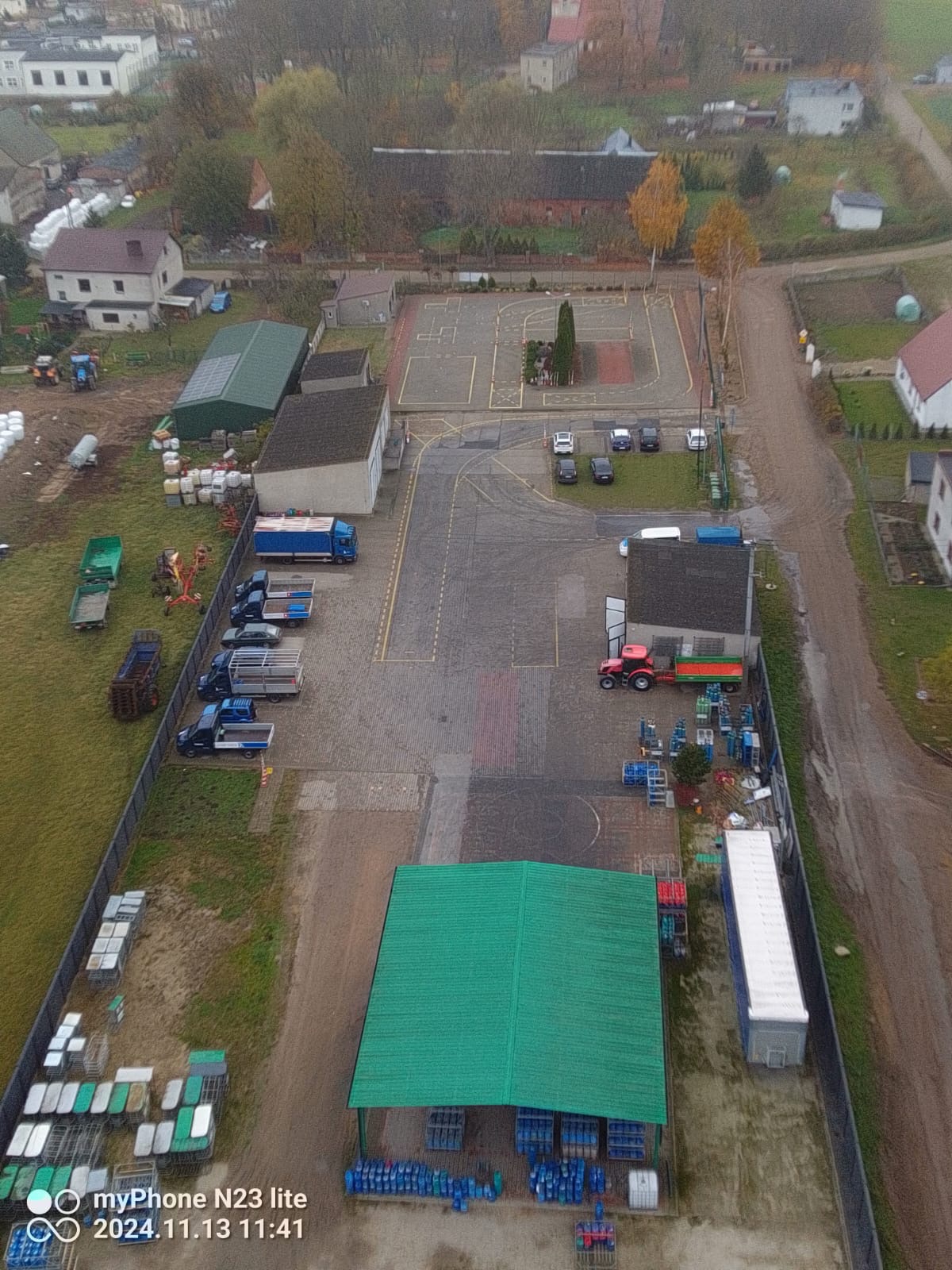 Widok z góry na plac manewrowy z samochodami osobowymi i ciężarowymi, budynki gospodarcze z zielonymi dachami, boisko z namalowanymi liniami i elementami infrastruktury, w tle zabudowa wiejska.