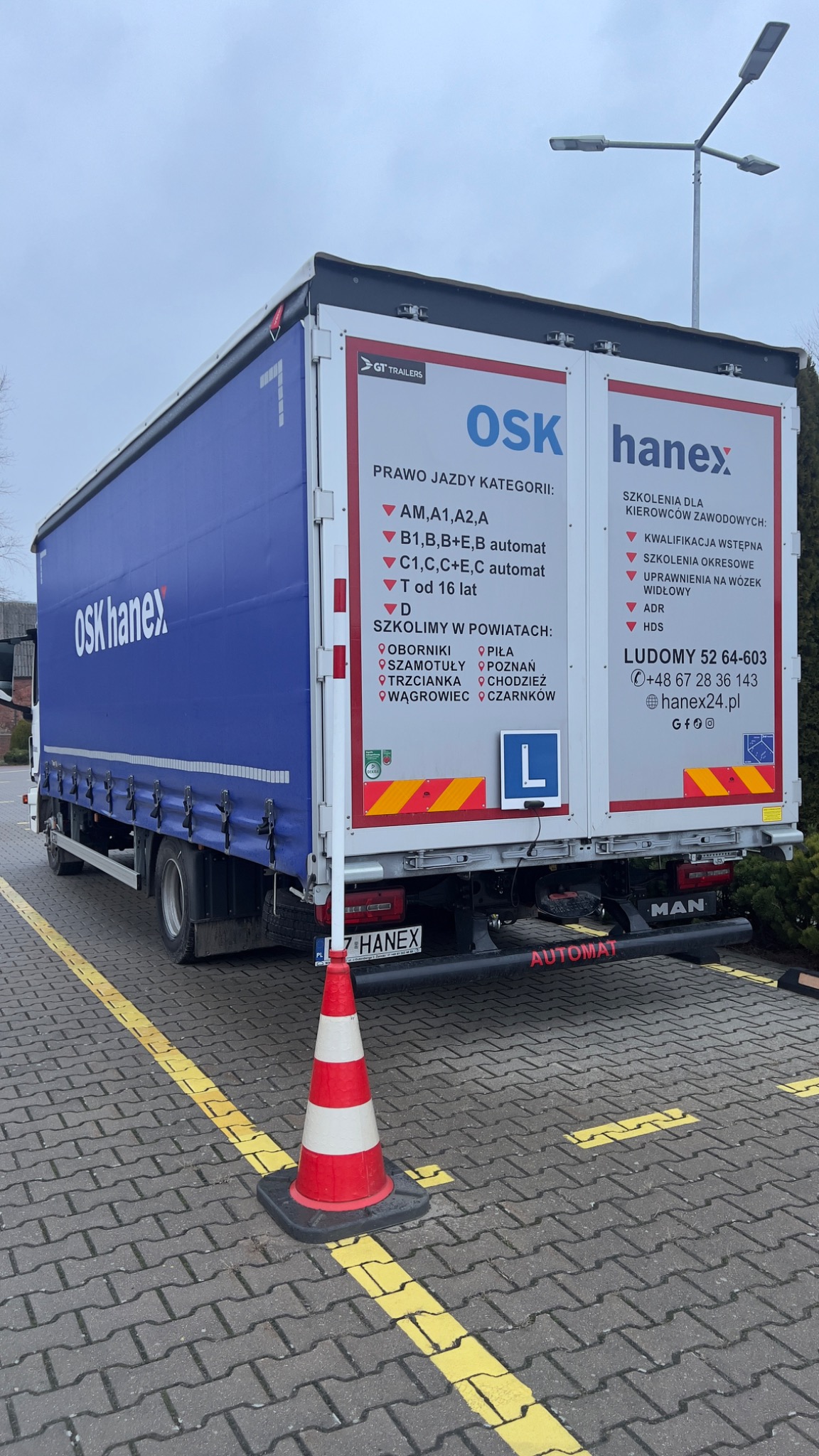Ciężarówka szkoły jazdy OSK Hanex z informacjami o kategoriach prawa jazdy i lokalizacjach szkoleń, zaparkowana na parkingu z żółtymi liniami, obok stoi pachołek drogowy.