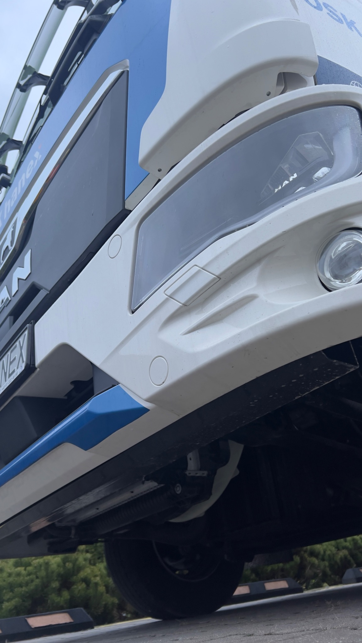 Niski kąt ujęcia na biało-niebieską kabinę ciężarówki MAN z widocznym reflektorem, czujnikami parkowania i fragmentem tablicy rejestracyjnej.