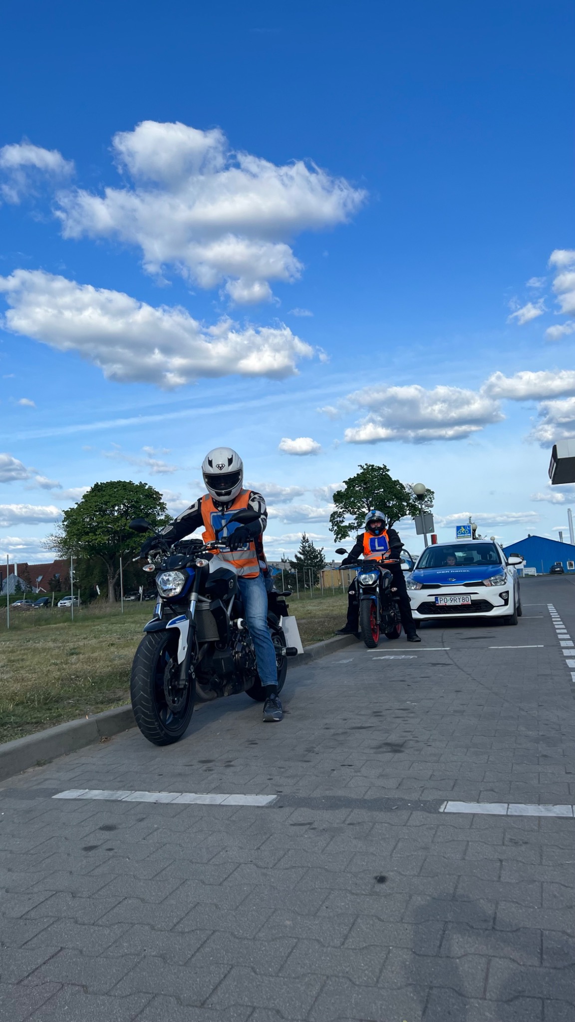 Dwaj kursanci na motocyklach w kamizelkach odblaskowych i kaskach, obok samochód oznaczony jako pojazd do nauki jazdy, na tle błękitnego nieba z chmurami.