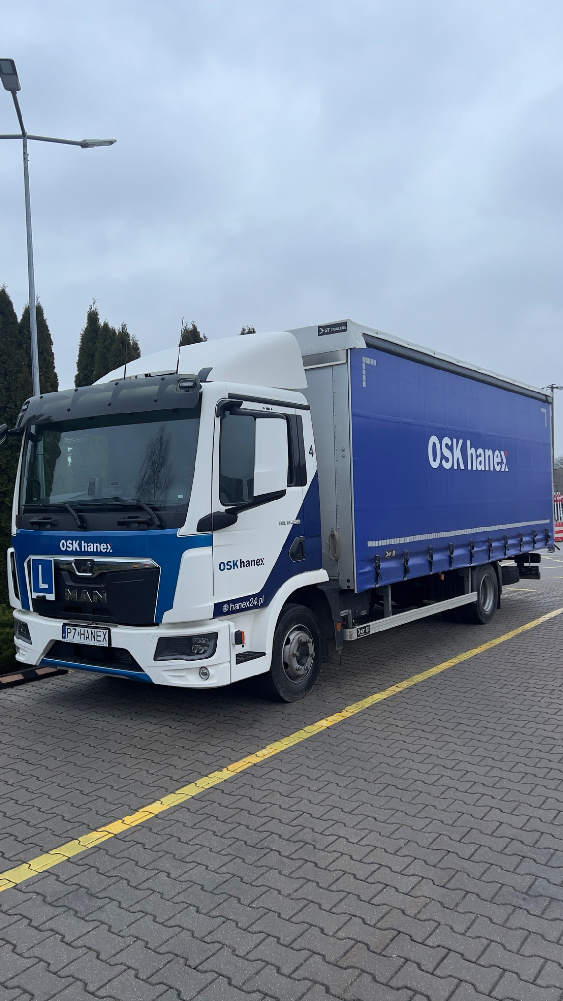 Ciężarówka MAN z oznakowaniem szkoły jazdy OSK Hanex, zaparkowana na brukowanym parkingu z żółtą linią oddzielającą miejsca postojowe.