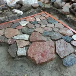 Układanie nieregularnych kamieni w ziemi, obramowanie z czerwonej cegły, widoczne przygotowane kamienie i wykop