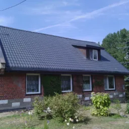 Dom z nowym, ciemnoszarym dachem z dachówki ceramicznej, widoczne okno dachowe, fragment elewacji z czerwonej cegły i ogród z krzewami.