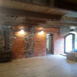 Surowe wnętrze z ceglaną ścianą, fragmentami kamiennej okładziny, drewnianym stropem, piecem wolnostojącym i panelami podłogowymi w odcieniach drewna. Widoczne drzwi łukowe i okno z widokiem...