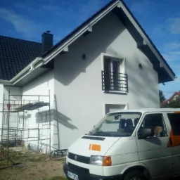 Świeżo pomalowany dom jednorodzinny w jasnych barwach, z rusztowaniem i samochodem dostawczym firmy malarskiej na pierwszym planie, widoczne niedociągnięcia w postaci plam po farbie na elewacji.