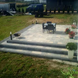 Nowoczesny taras z szarych płyt, z trzema stopniami obłożonymi kostką, zestaw mebli ogrodowych i rośliny w donicach na tle zadbanego trawnika oraz zabudowań gospodarczych.