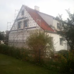 Dom jednorodzinny w trakcie ocieplania elewacji, widoczne rusztowania i płyty izolacyjne mocowane do ściany, czerwona dachówka, pochmurne niebo w tle.