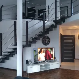 Nowoczesne schody wewnętrzne z metalową balustradą i drewnianymi stopniami w przestronnym salonie z telewizorem i dekoracyjnym zegarem na ścianie.