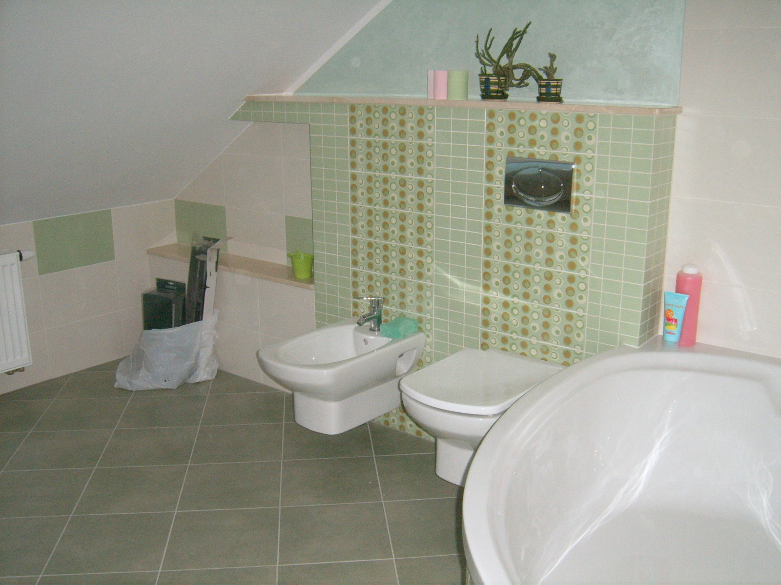 Ukończona łazienka na poddaszu z zielono-beżowymi płytkami w kropki, białą wanną, bidetem i toaletą; na półce dekoracje i kosmetyki.