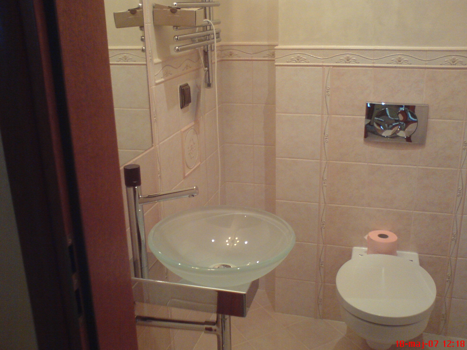 Jasna łazienka z umywalką ze szkła, chromowaną armaturą i beżowymi płytkami z dekoracyjnym paskiem pionowym. Na spłuczce toalety leży rolka papieru toaletowego w kolorze różowym.