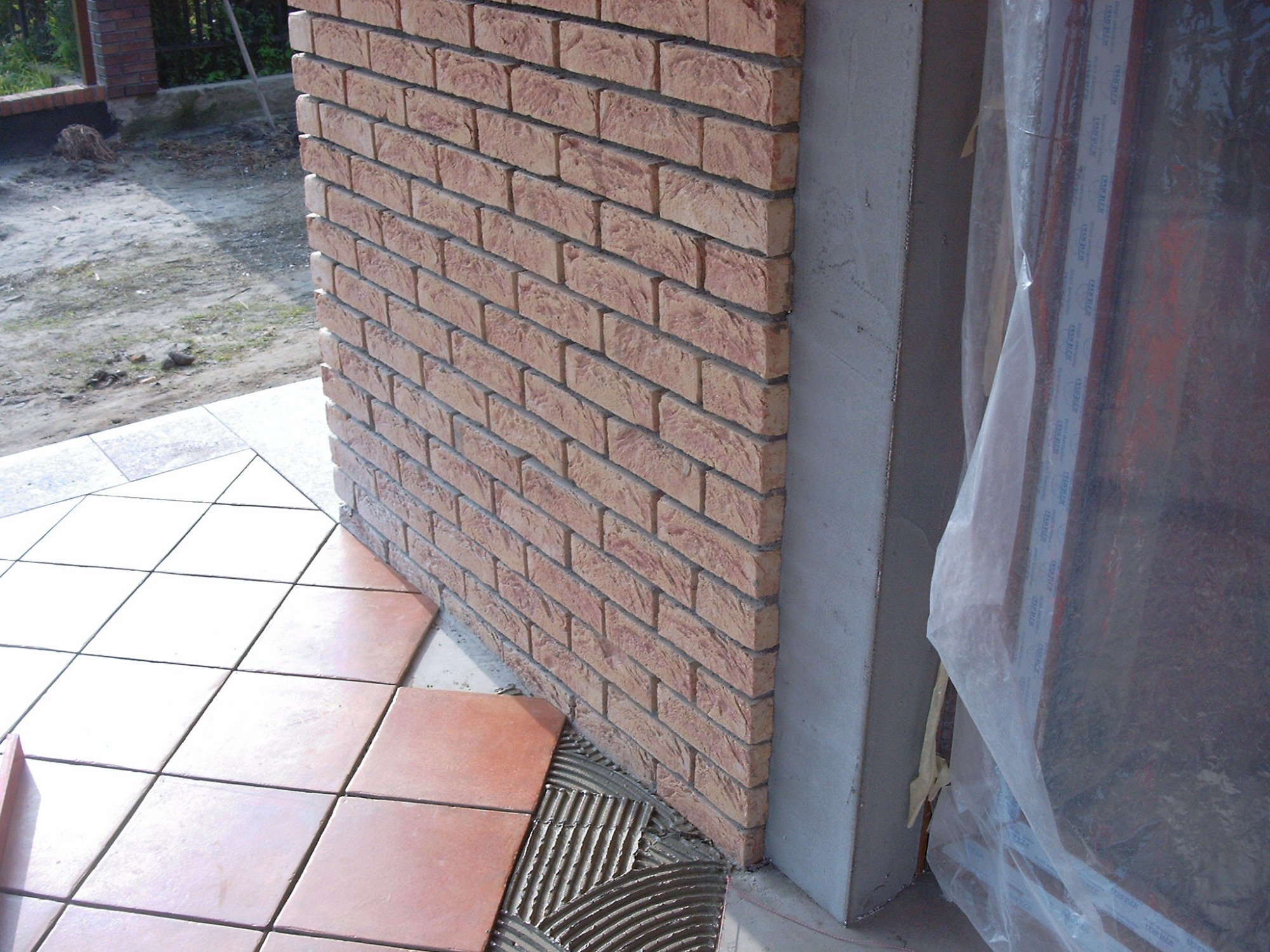 Fragment ściany wykończonej cegłą klinkierową, z częściowo ułożoną podłogą z terakoty, widoczne ślady zaprawy klejowej, zabezpieczona folią okienna rama.