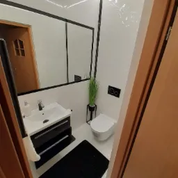 Nowoczesna, biało-czarna łazienka z podświetlanym sufitem, minimalistyczną umywalką z szafką, toaletą i czarnym dywanikiem, widziana z perspektywy otwartych drzwi.