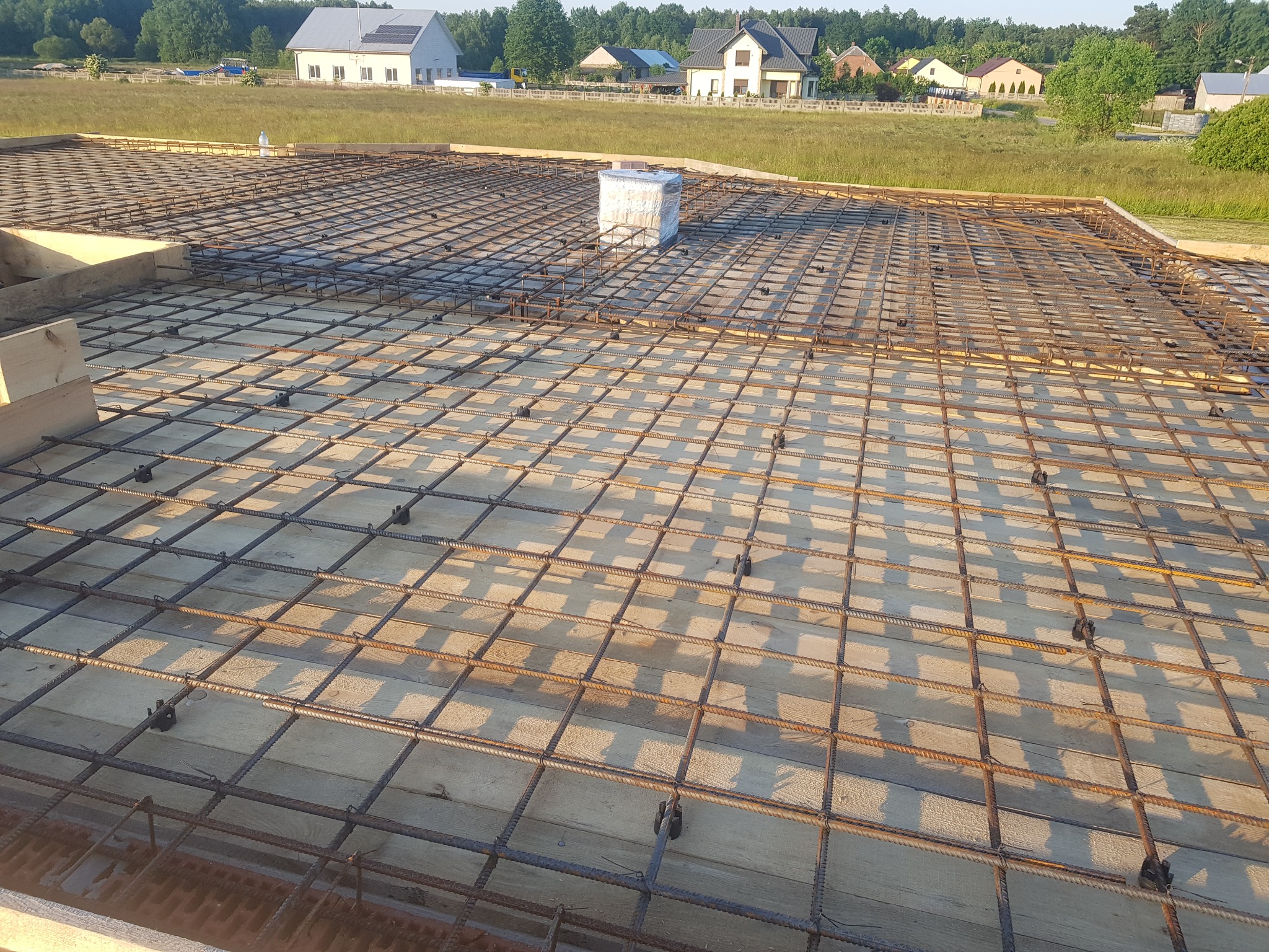 Siatka zbrojeniowa ułożona na drewnianym szalunku, przygotowana do wylania betonu, z widocznym opakowaniem materiałów budowlanych w tle krajobrazu wiejskiego.