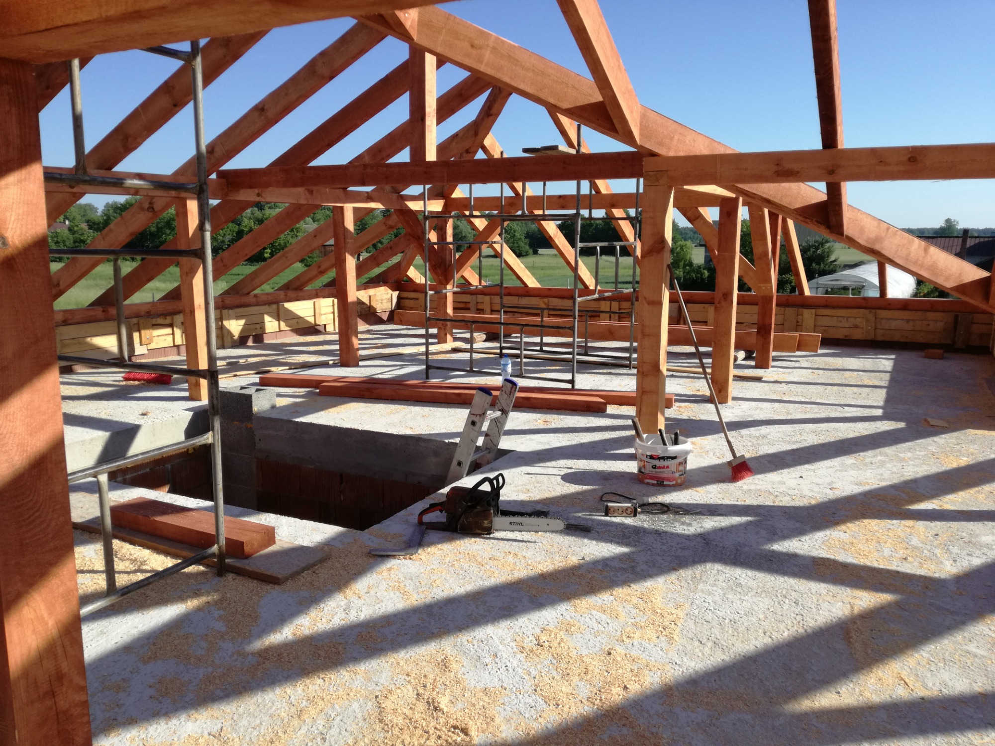 Konstrukcja drewniana domu w trakcie budowy, widoczne krokwie, słupy, podłoga pokryta wiórami, drabina, piła spalinowa Stihl i wiadro z narzędziami.