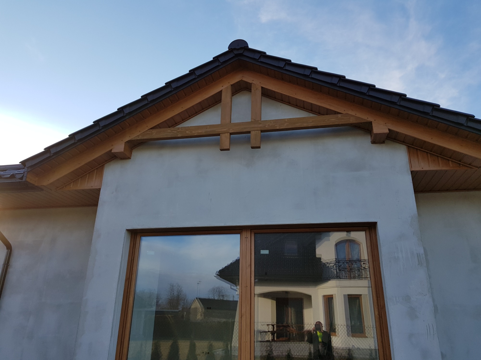 Surowy stan elewacji domu z widoczną drewnianą konstrukcją dachu i dużym oknem odbijającym sąsiedni budynek.