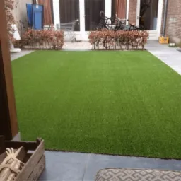 Równy trawnik z zielonej, sztucznej trawy na betonowym podłożu, z niskimi krzewami w tle i drewnianą skrzynią z dekoracjami w rogu.