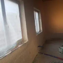 Wnętrze w trakcie prac: ściany z nałożoną, nierówną warstwą zaprawy, okna zabezpieczone folią malarską, widoczne rury i wiadro, sufit z betonowych płyt.