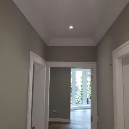 Korytarz po remoncie z szarymi ścianami, białymi futrynami drzwi i ozdobnym białym gzymsem sufitowym oraz oświetleniem punktowym.