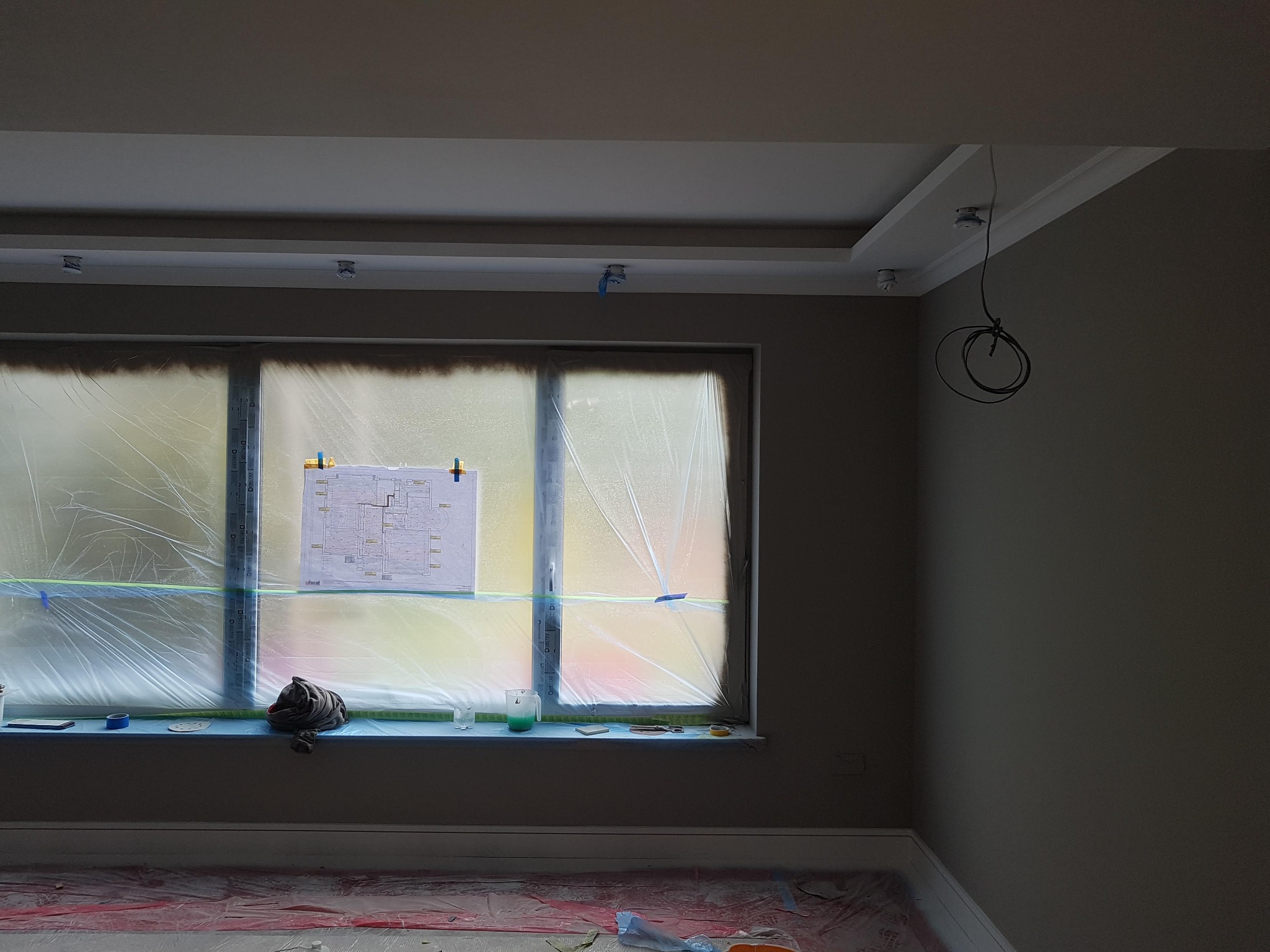 Pokój w trakcie remontu: okno zabezpieczone folią, na parapecie narzędzia i plan, podłoga zakryta folią ochronną, widoczne przygotowane ściany i sufit z oświetleniem.