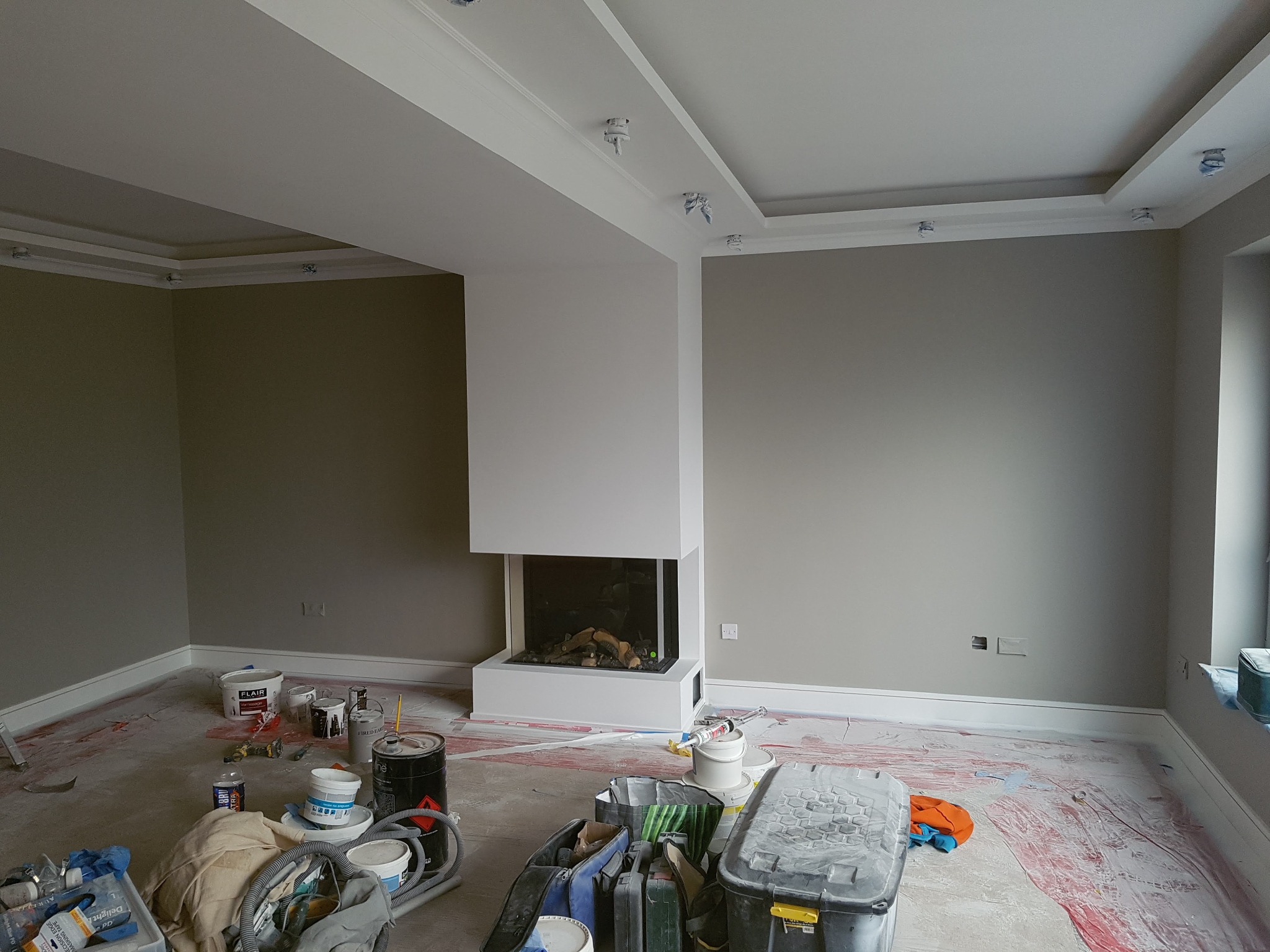 Pokój w trakcie remontu: ściany pomalowane na szaro i biało, widoczny kominek, narzędzia i farby na podłodze zabezpieczonej folią.