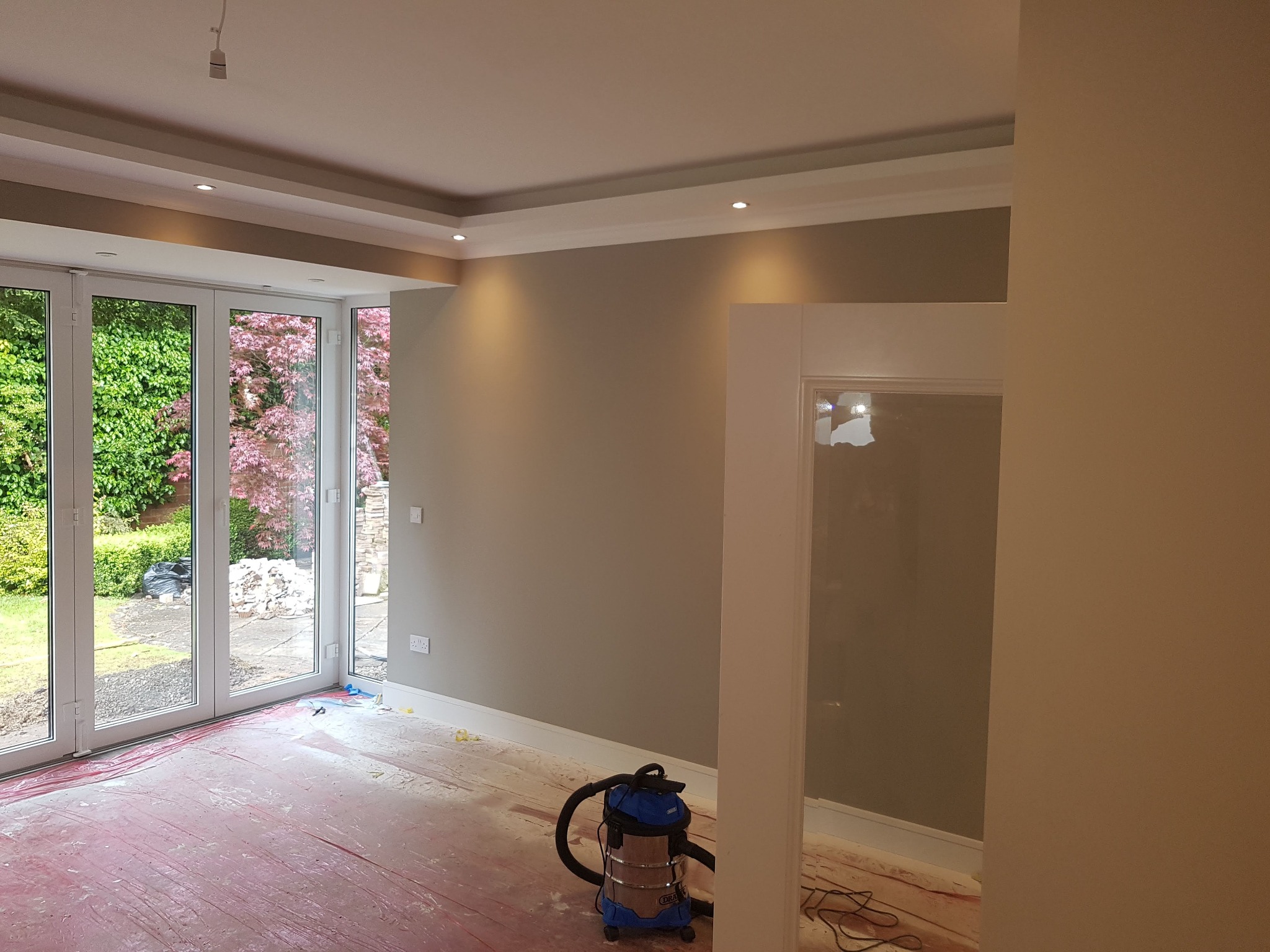 Pokój po malowaniu ścian na szaro, z widocznymi listwami przypodłogowymi, osłoniętą podłogą i odkurzaczem przemysłowym, duże okno balkonowe z widokiem na zieleń.