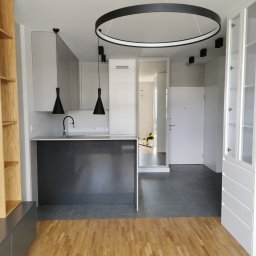 TSE Remonty - Nowoczesna kuchnia z wyspą, szare szafki, czarne lampy wiszące i okrągła lampa sufitowa, drewniana podłoga i szara podłoga w kuchni, widok na przedpokój z białymi drzwiami.